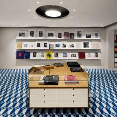 Innenraum eines modernen Showrooms mit XAL SASSO PRO 80 adjustable flush round recessed Downlights in der Decke. Minimalistische Einrichtung mit Bücherregalen, Ausstellungsfläche und auffälligem blau-weißem Bodenmuster
